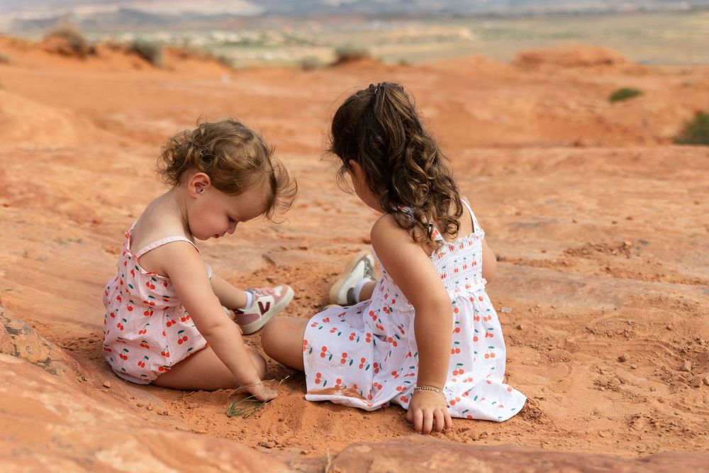 Two young girls in floral dresses sitting on red desert sand, playing together with small rocks outdoors.