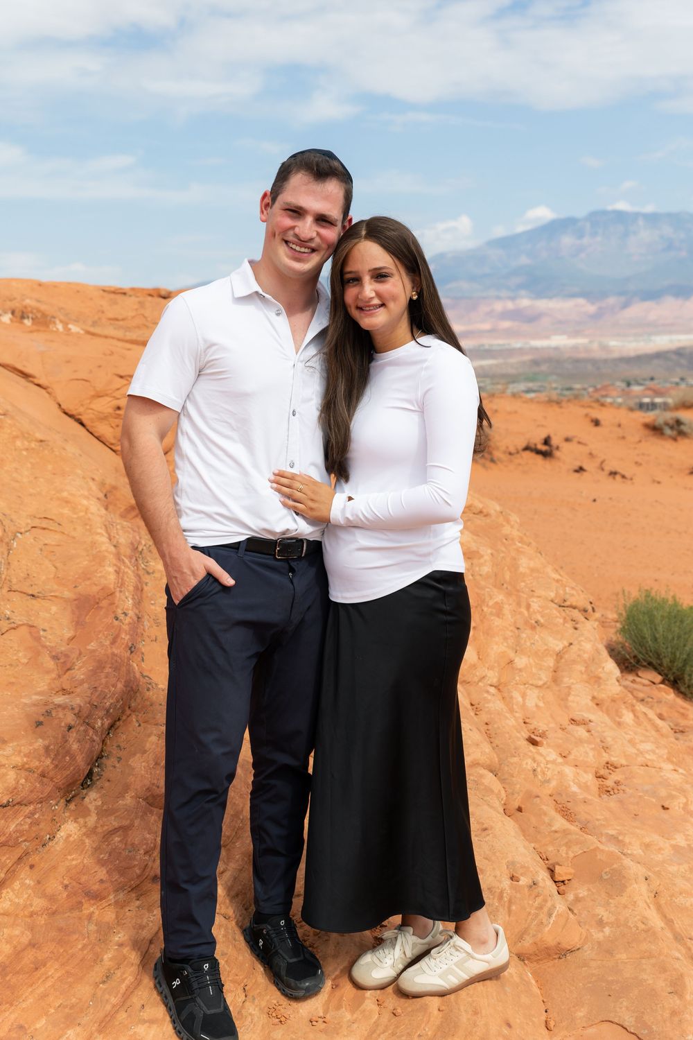 Couple posing together on red rock formations in a desert landscape, woman in white top and black skirt, man in white shirt.
