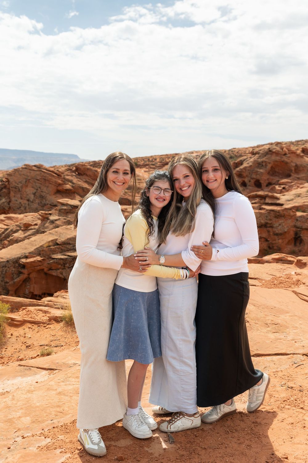 Four young women posing together outdoors at a red rock canyon landscape, smiling and embracing.