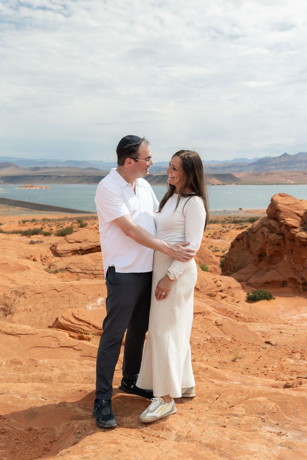 Couple embracing on red rock desert landscape with Lake Powell and mountains in background on cloudy day.
