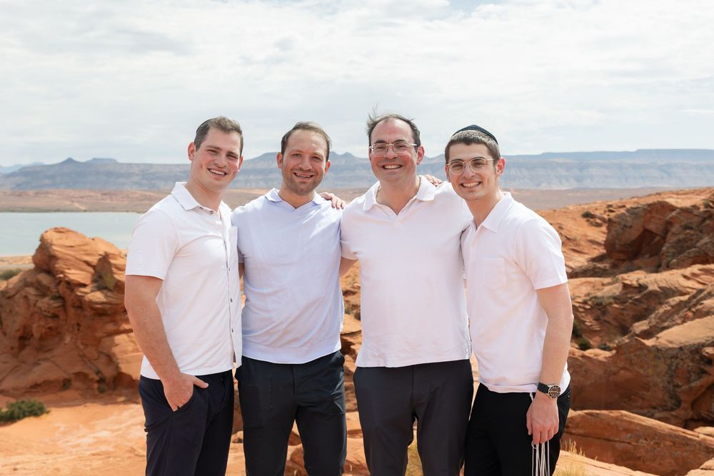 Four men in white polo shirts smiling together at a red rock canyon overlook with a lake and blue sky behind them.