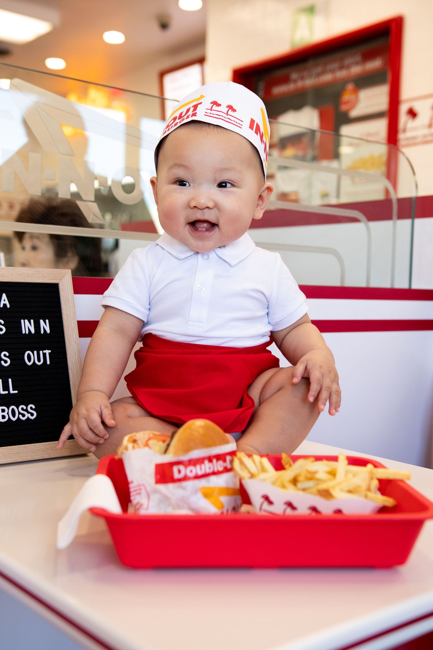 Adorable baby in In-N-Out Burger uniform sitting on counter with Double-Double meal and fries in red tray.