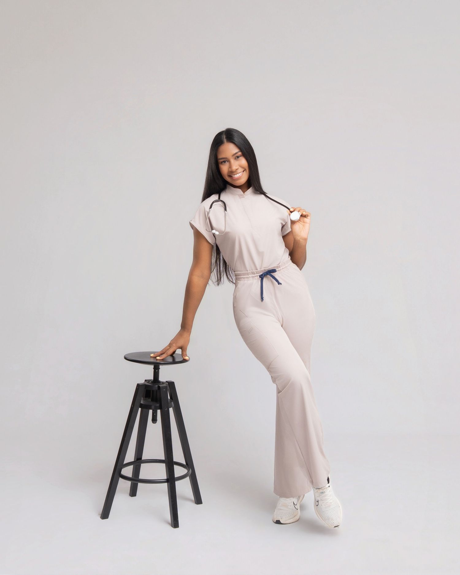 Woman in beige medical scrub jumpsuit poses with stethoscope beside black stool on gray background.