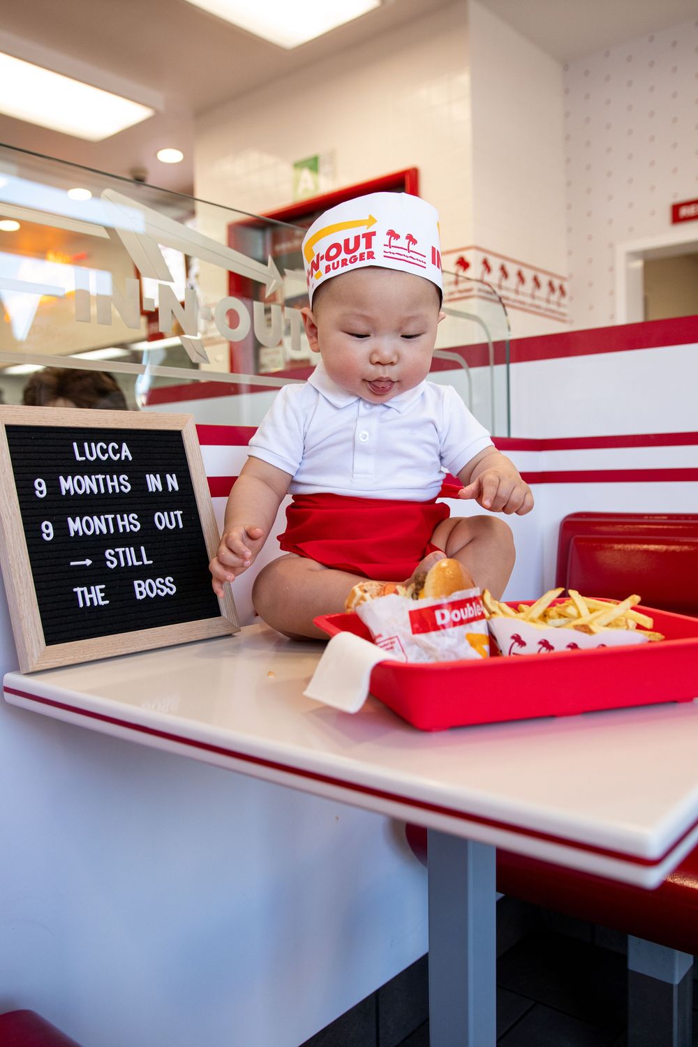 Baby dressed as In-N-Out Burger employee sits on restaurant table with fries, sign reads Lucca 9 months old.