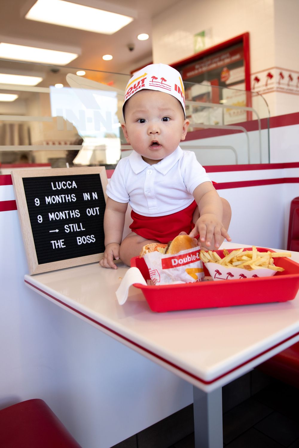Baby Lucca in In-N-Out uniform sits at restaurant booth with burger and fries tray, celebrating 9 months old.