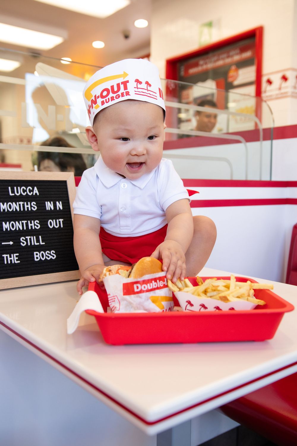 Adorable baby dressed in In-N-Out Burger uniform sitting at restaurant counter with food tray and milestone sign.