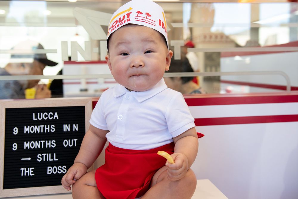 Baby Lucca in In-N-Out Burger outfit sits with fries at 8 months, wearing a paper hat and red shorts.