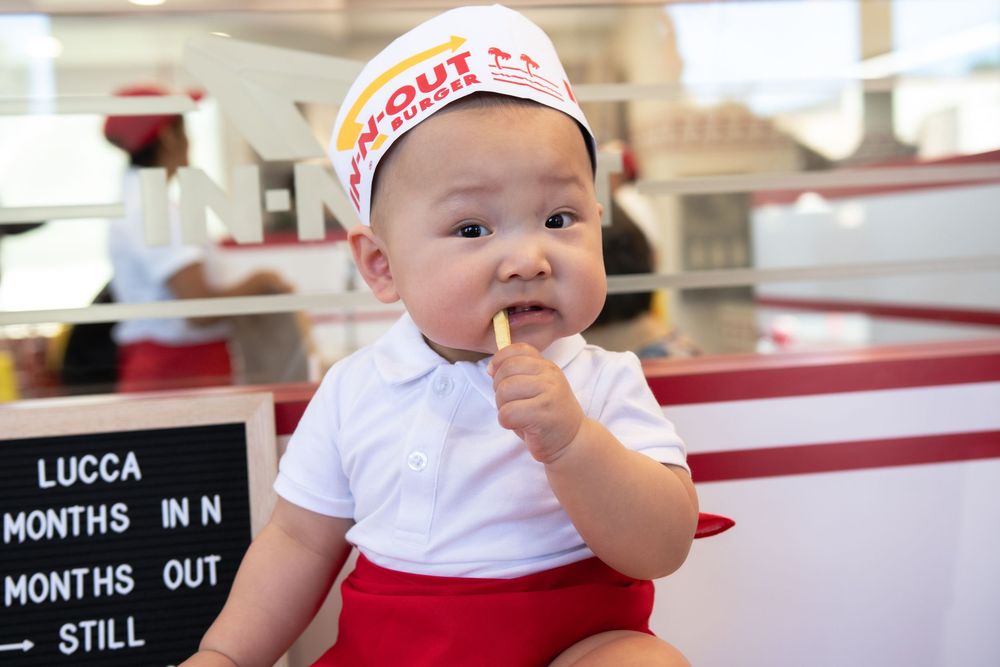 Baby dressed in In-N-Out Burger uniform holding a french fry, sitting next to a chalkboard sign.