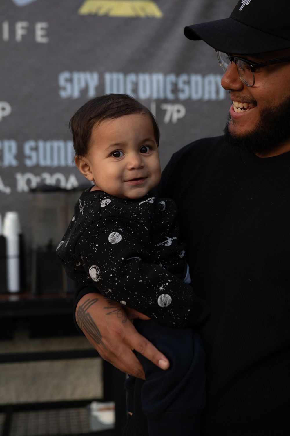 A smiling man in a black cap holds a cute baby wearing a dark space-themed outfit at an indoor event.