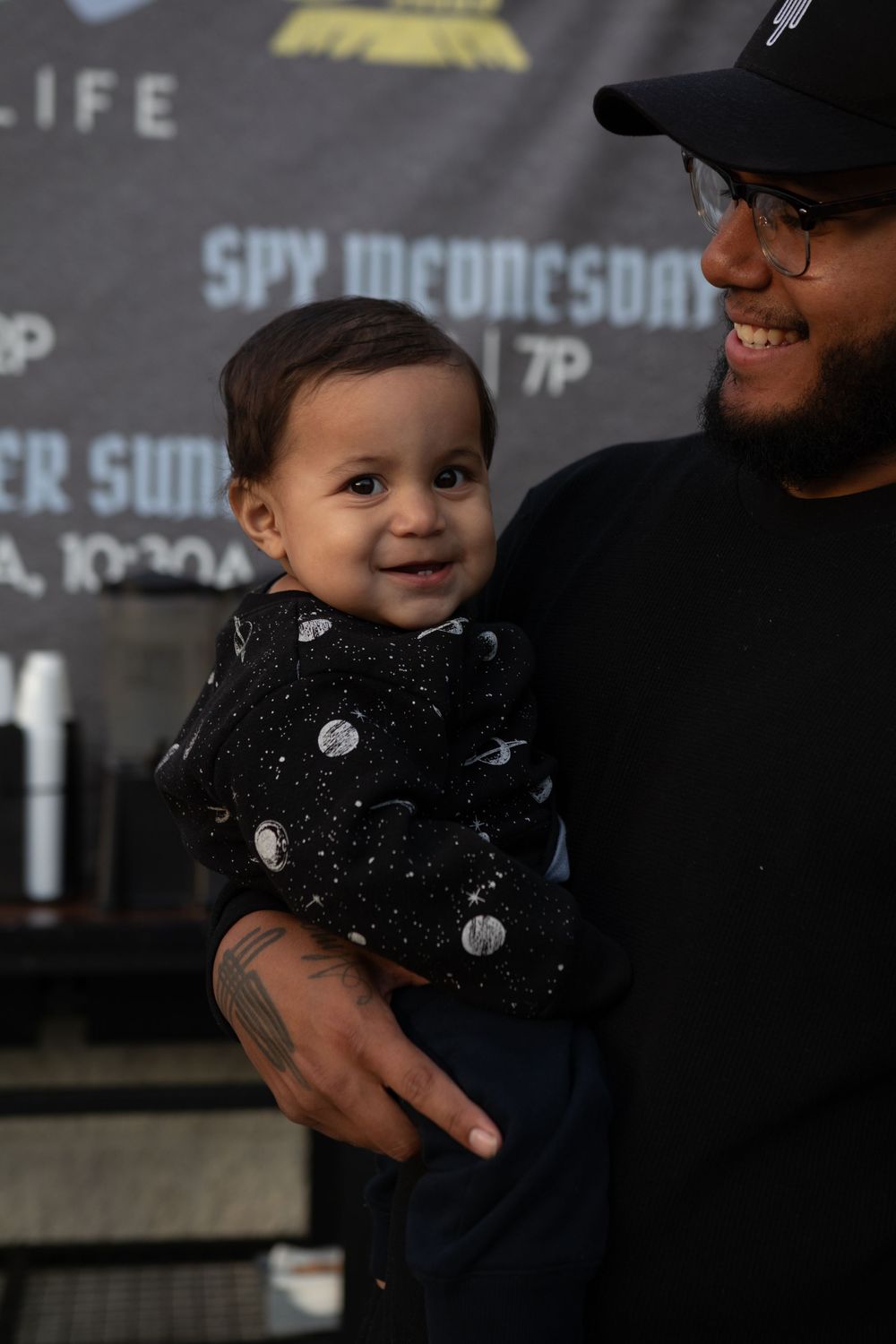 A smiling baby in a space-print outfit being held by a man in a black cap and shirt at an event.