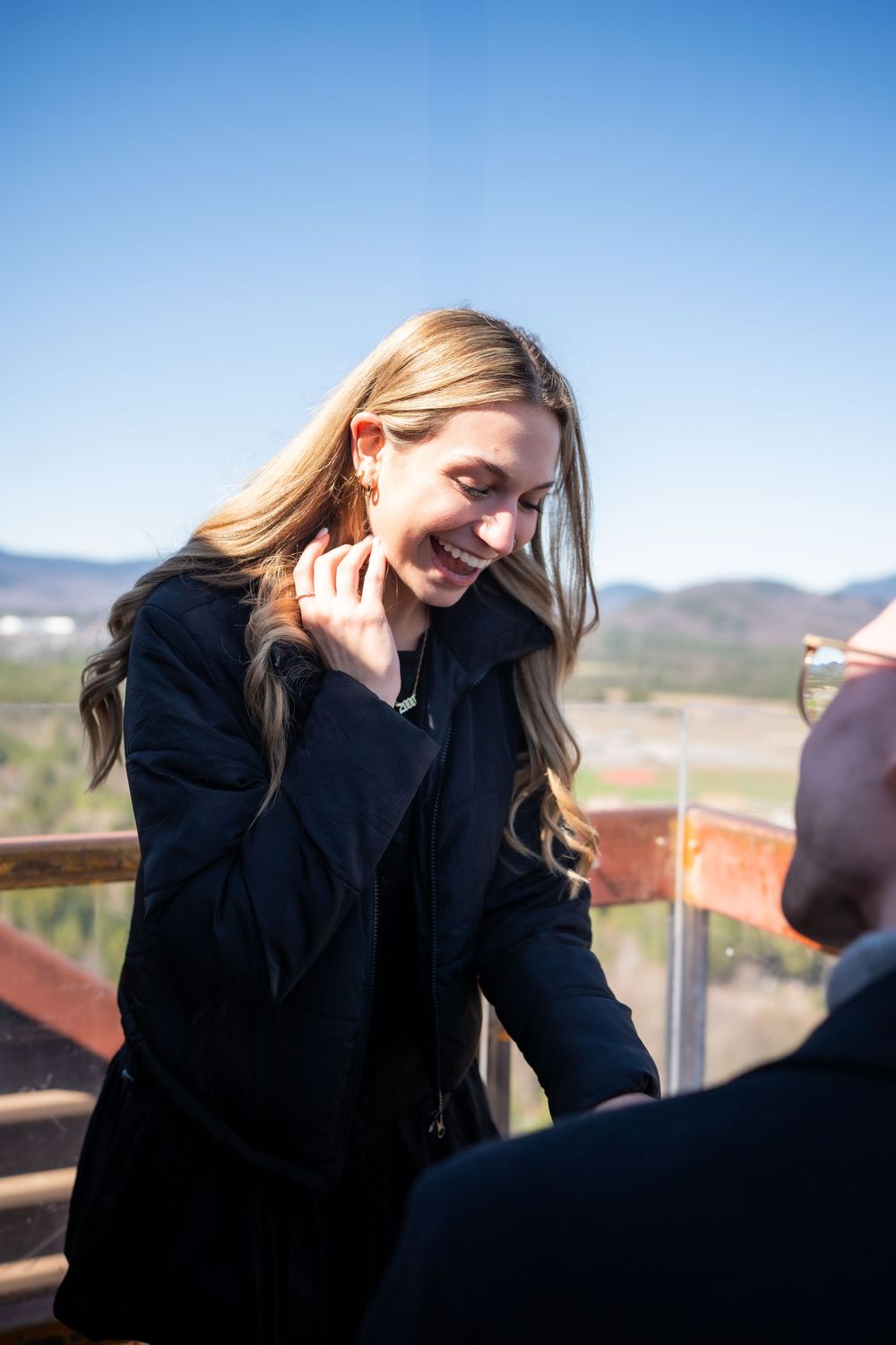 Smiling blonde woman in black coat on outdoor overlook with mountain landscape and blue sky background.