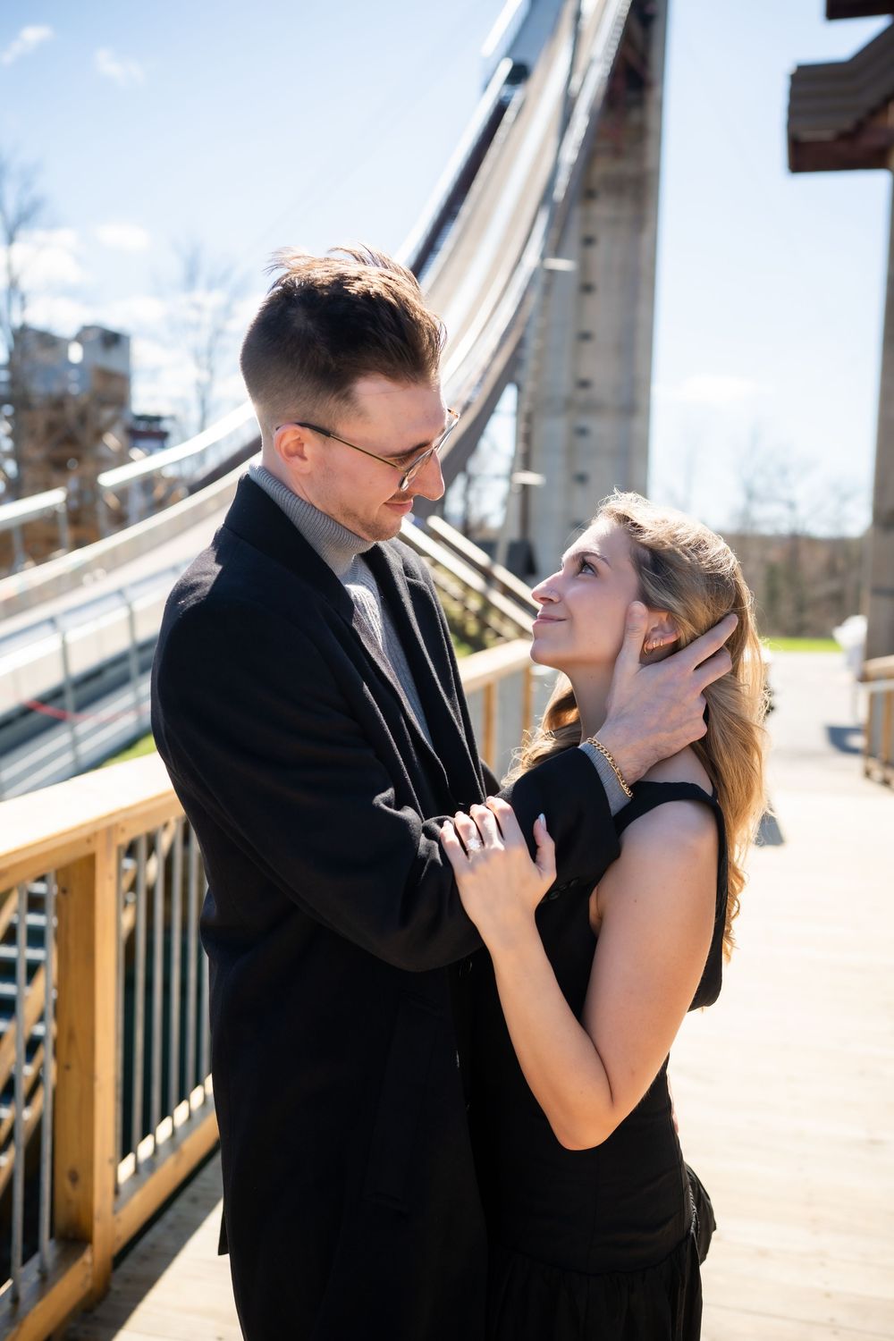A couple in black attire share a tender moment on a bridge, the man gently holding the woman's face as she gazes up at him.