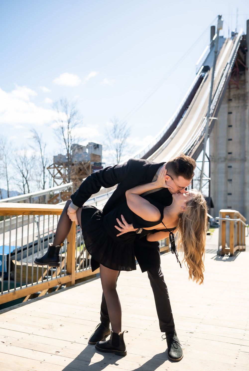 Couple in black outfits sharing a romantic dip kiss on a wooden deck with a ski jump structure behind them.