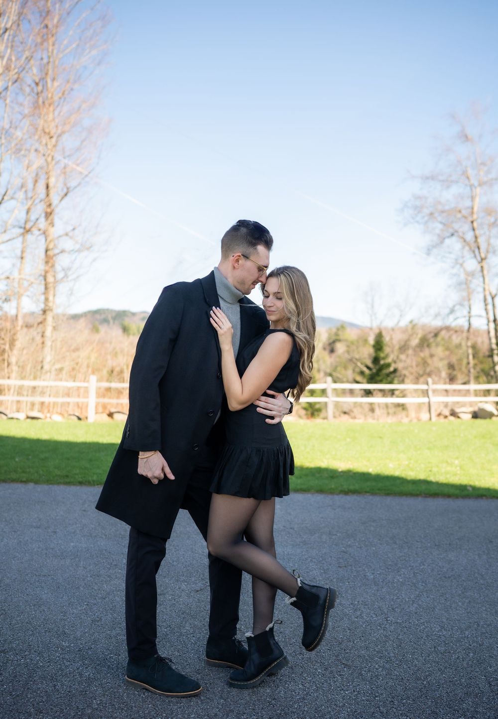 Couple in matching black outfits sharing a romantic moment outdoors with mountains and white fence backdrop.