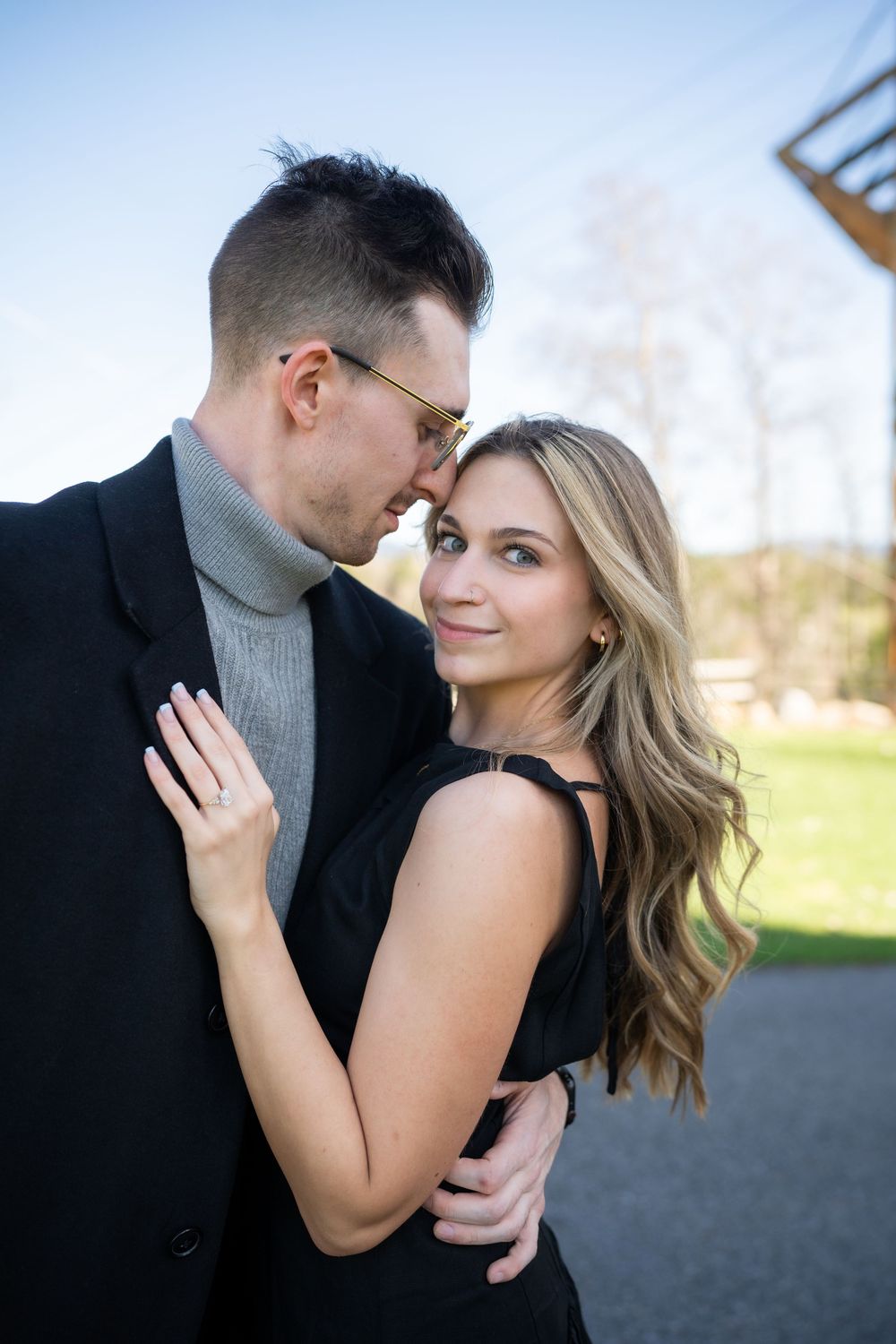 A couple in black outfits share a romantic moment outdoors, woman smiling at camera while man leans in close.