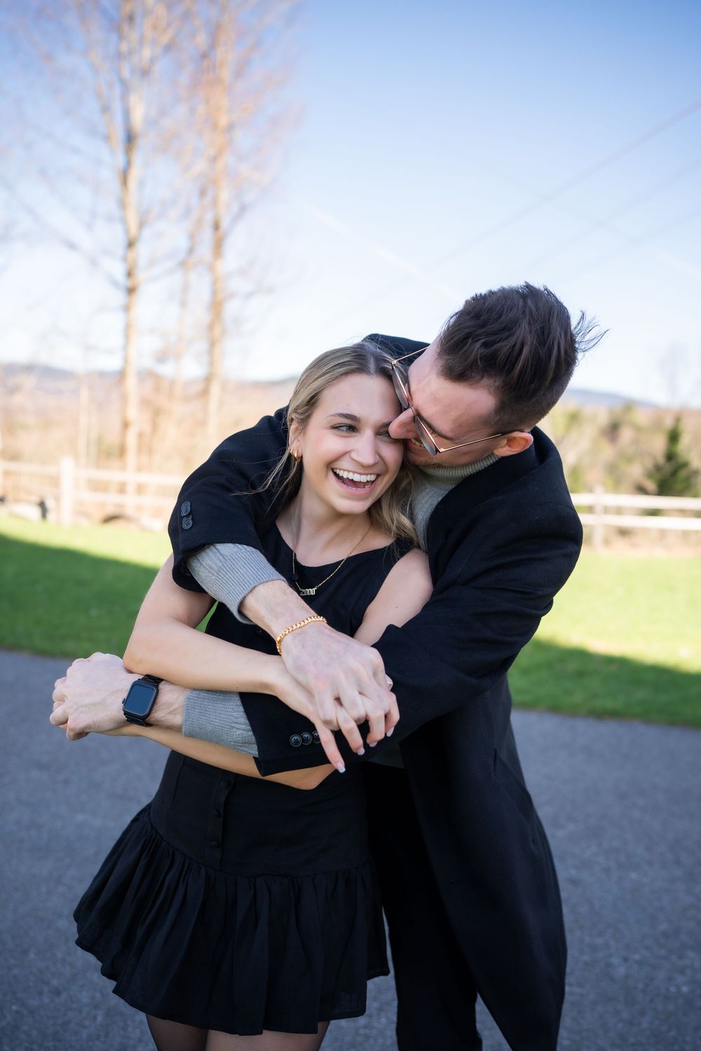 Happy couple in black outfits embracing outdoors, woman showing engagement ring, laughing joyfully on a sunny day.