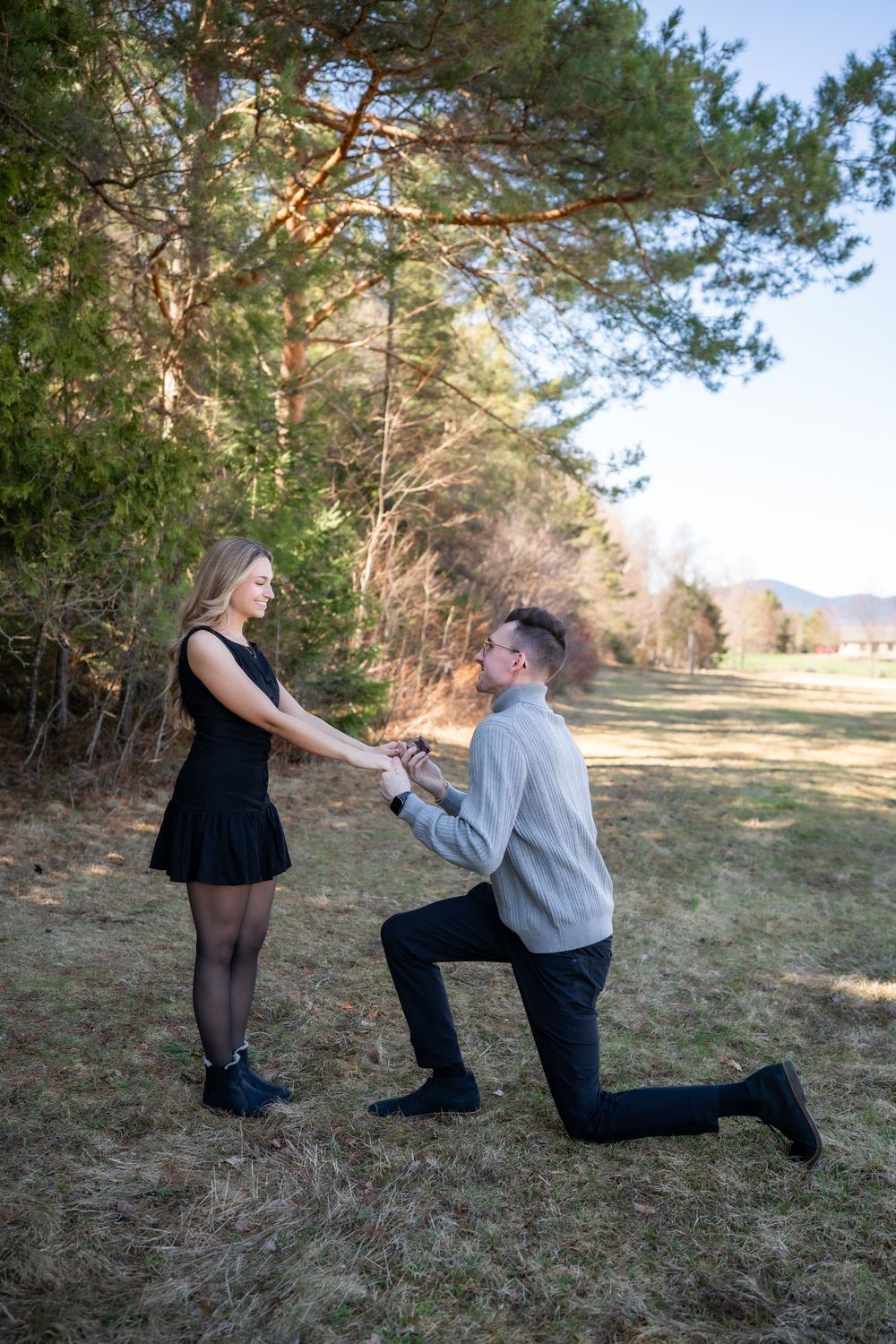 Man proposes on one knee to woman in black dress outdoors, holding hands in a sunny forest clearing.