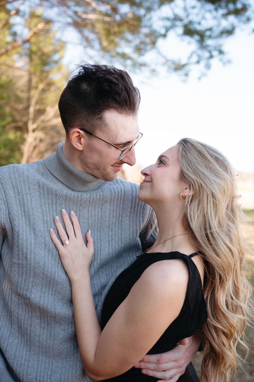 A couple gazes lovingly at each other outdoors, man in gray turtleneck with glasses, woman in black top with long blonde hair.