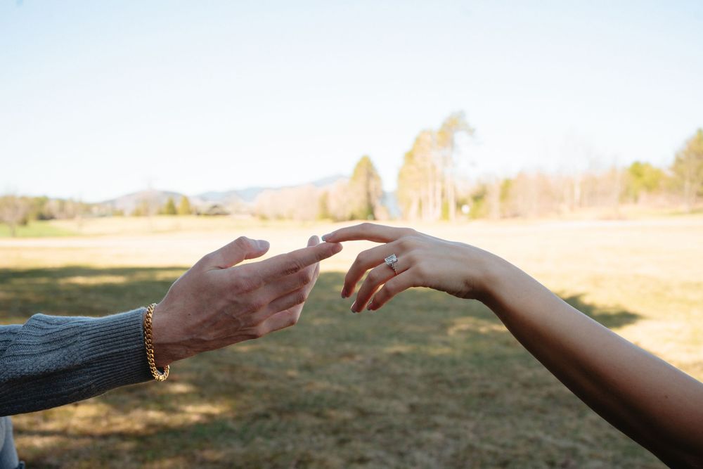Two hands reaching toward each other outdoors in a field, one wearing a ring, with mountains and trees in the background.