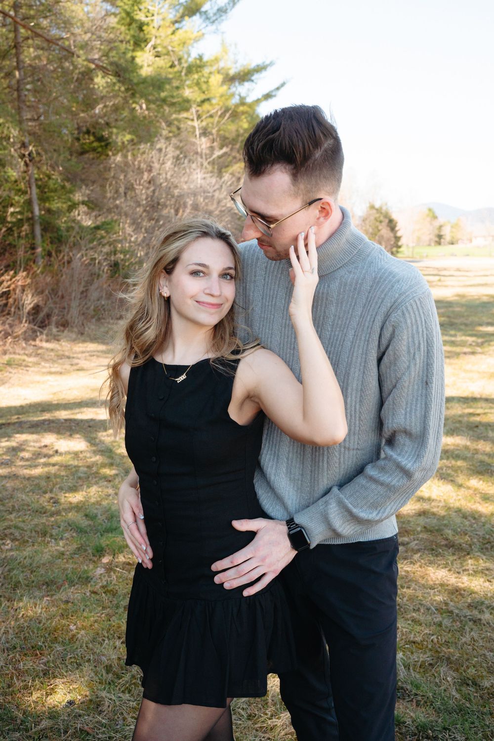 A couple poses outdoors, woman in black dress smiling, man in gray sweater embracing her with trees in background.