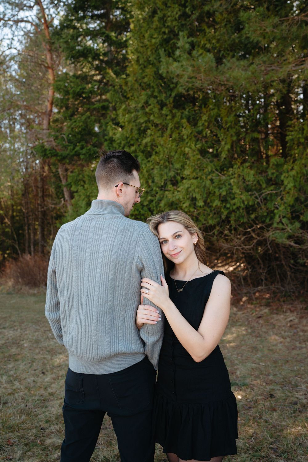 A smiling young woman in a black dress holds a man in a gray sweater outdoors near evergreen trees.