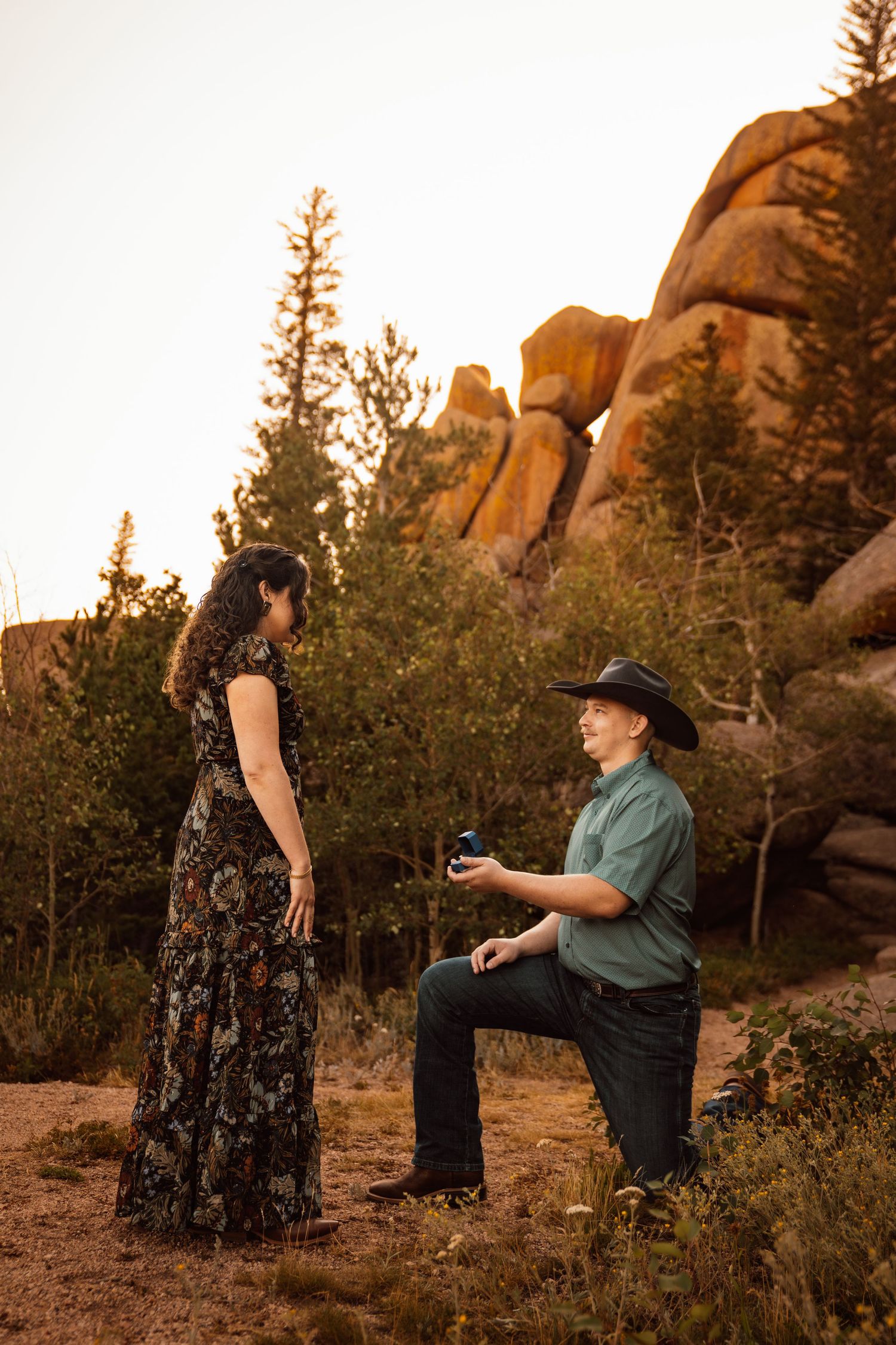 Man in cowboy hat proposes on one knee to woman in floral dress at golden hour near red rock formations.