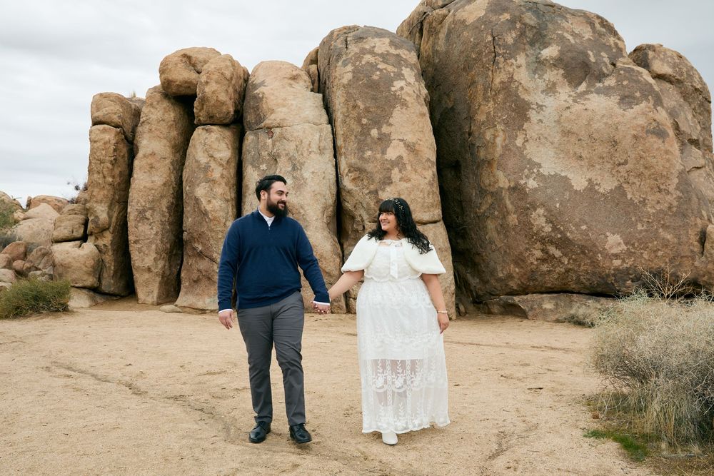 Couple holding hands walking among large desert boulders, man in navy sweater and woman in white lace dress, Joshua Tree.