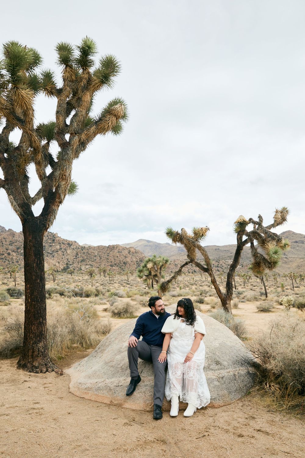 Couple sitting on a boulder among Joshua trees in the desert landscape of Joshua Tree National Park.