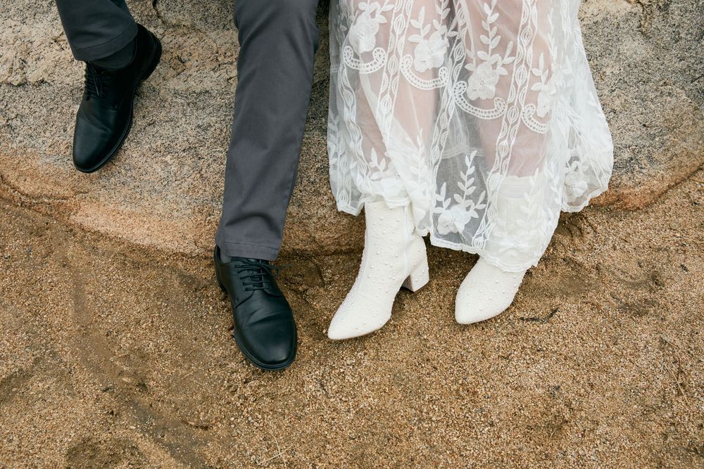 Close-up of wedding couple's feet, groom in black dress shoes and bride in white lace cowboy boots on sandy ground.