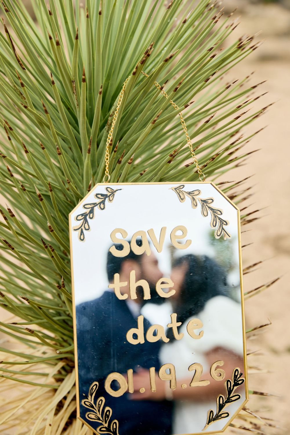 Save the date sign reading 01.19.26 with couple reflection, leaning against desert yucca plant on sandy ground.