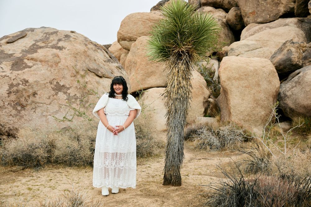 Woman in white lace dress standing beside a yucca tree with large boulders in Joshua Tree desert landscape.