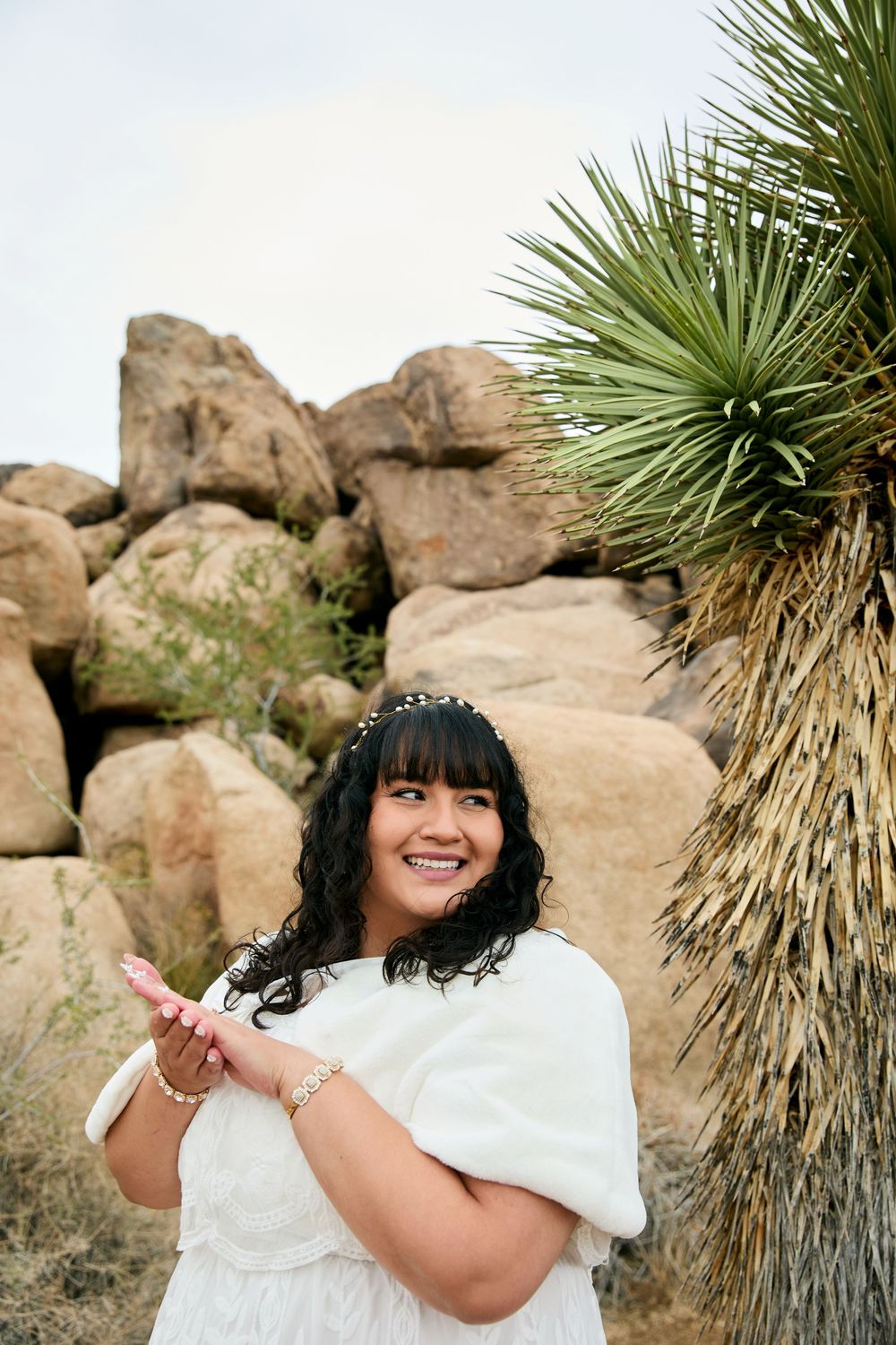 Smiling bride in white dress with floral headband poses near Joshua tree and rocky boulders.