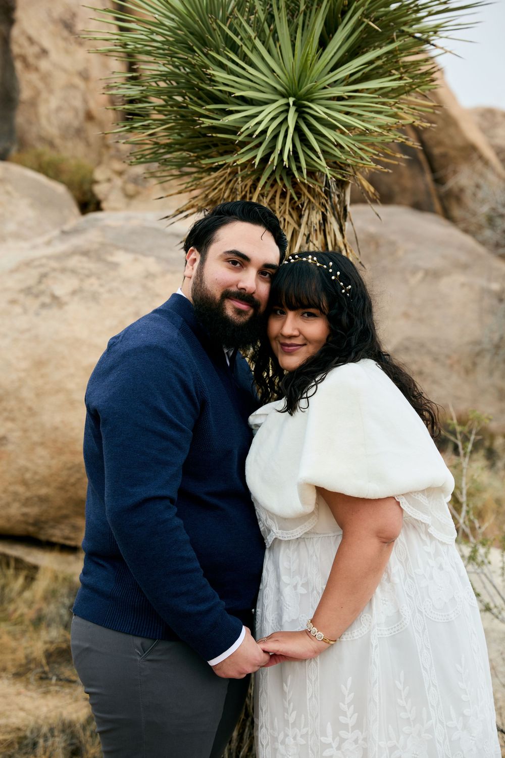 Couple posing together in front of yucca tree and boulders, woman in white dress and man in blue sweater.