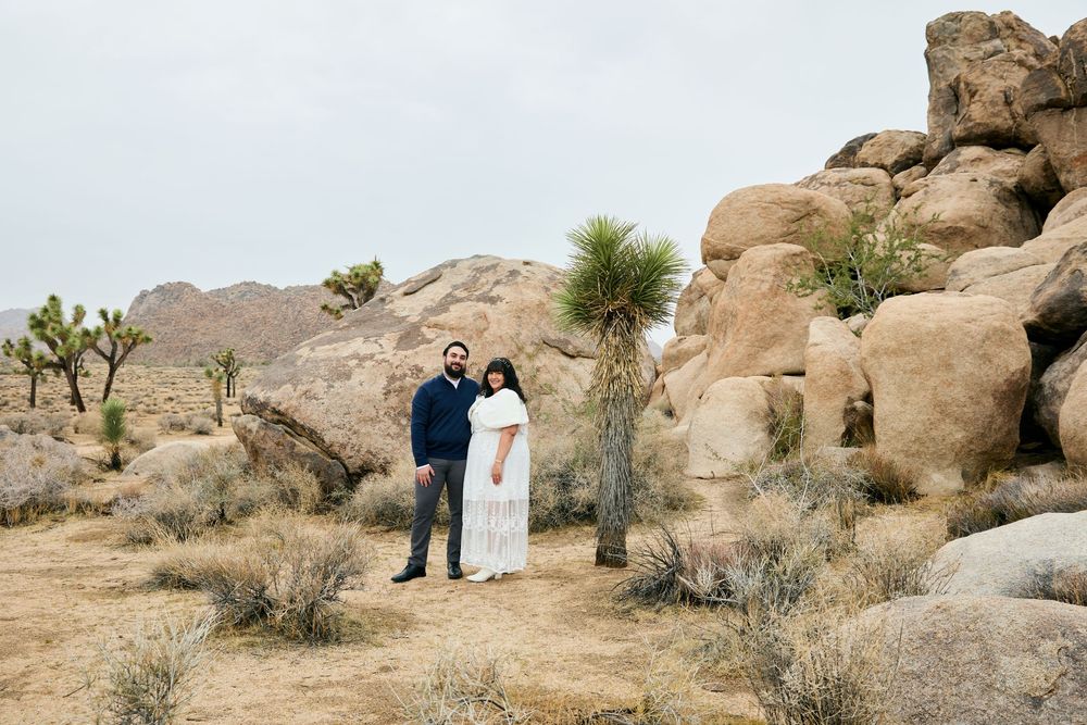 Couple stands together in Joshua Tree desert landscape with rocky boulders and cacti under overcast sky.