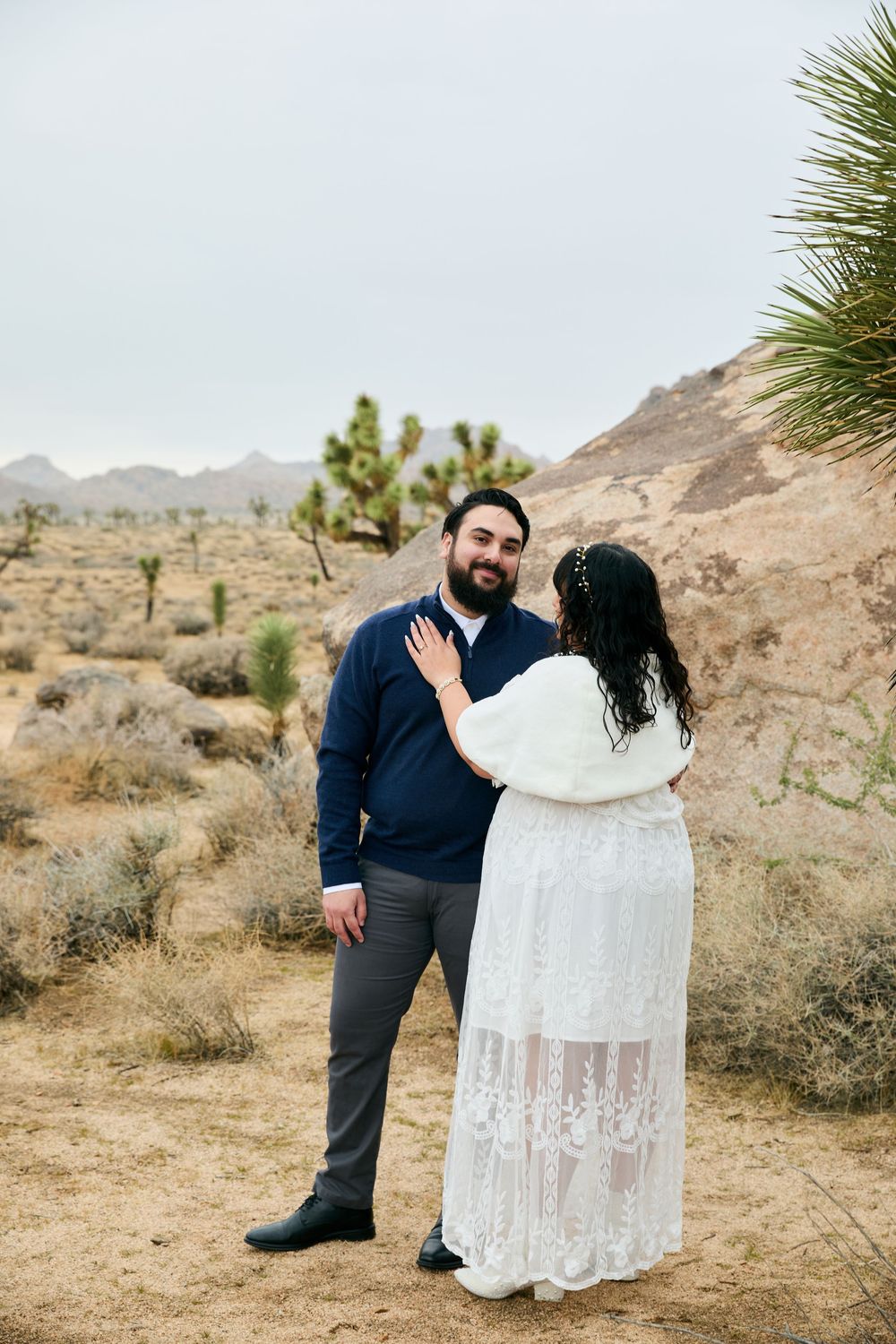 Couple in wedding attire sharing a romantic moment in Joshua Tree desert landscape with mountains in background.