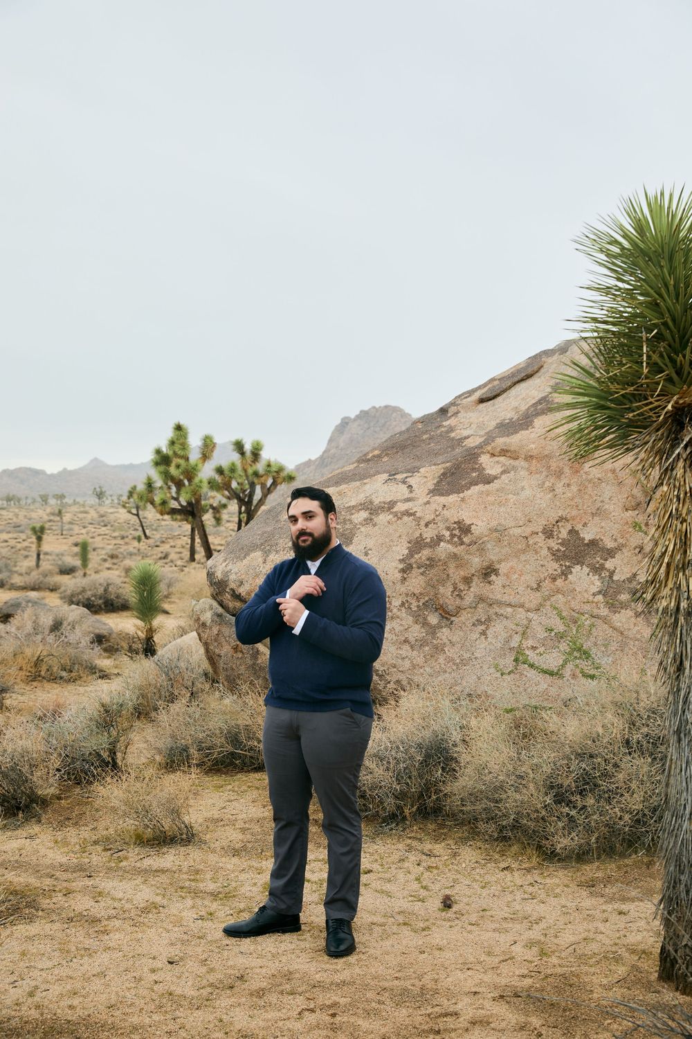 Bearded groom in navy sweater adjusts cufflinks among Joshua trees and boulders in desert landscape.