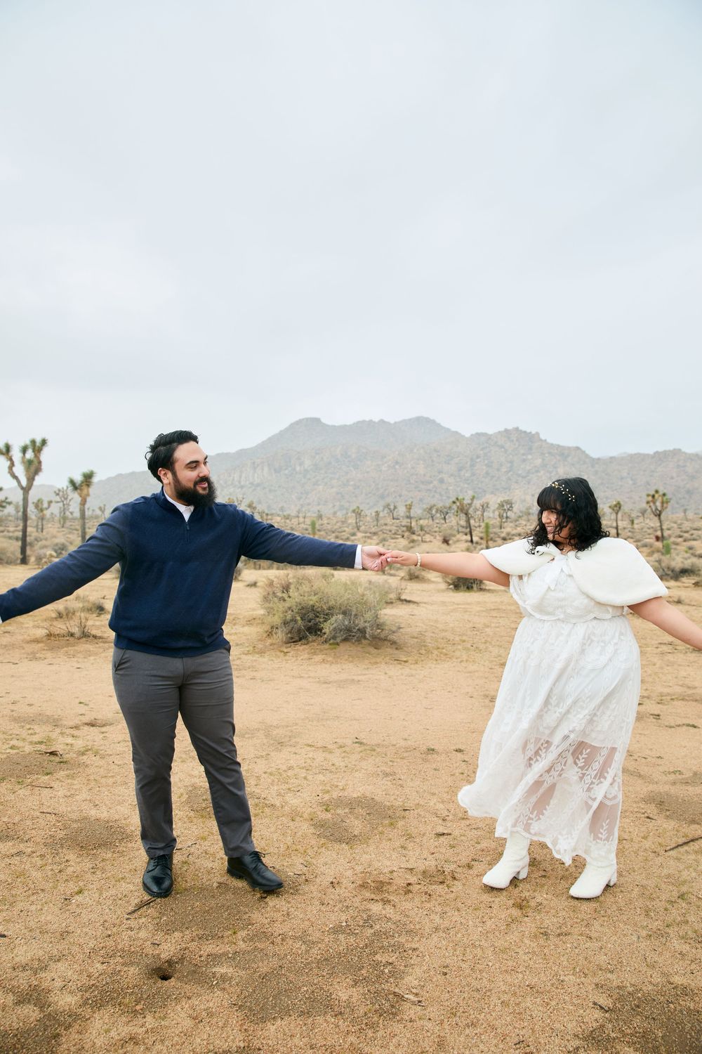 Couple dancing in Joshua Tree desert landscape, woman in white lace dress, man in navy sweater, mountains behind.