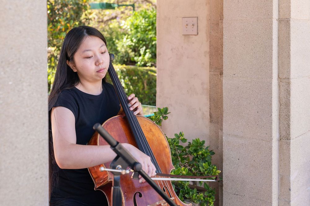 Young woman playing cello outdoors near stone columns, focused on her performance in natural light.