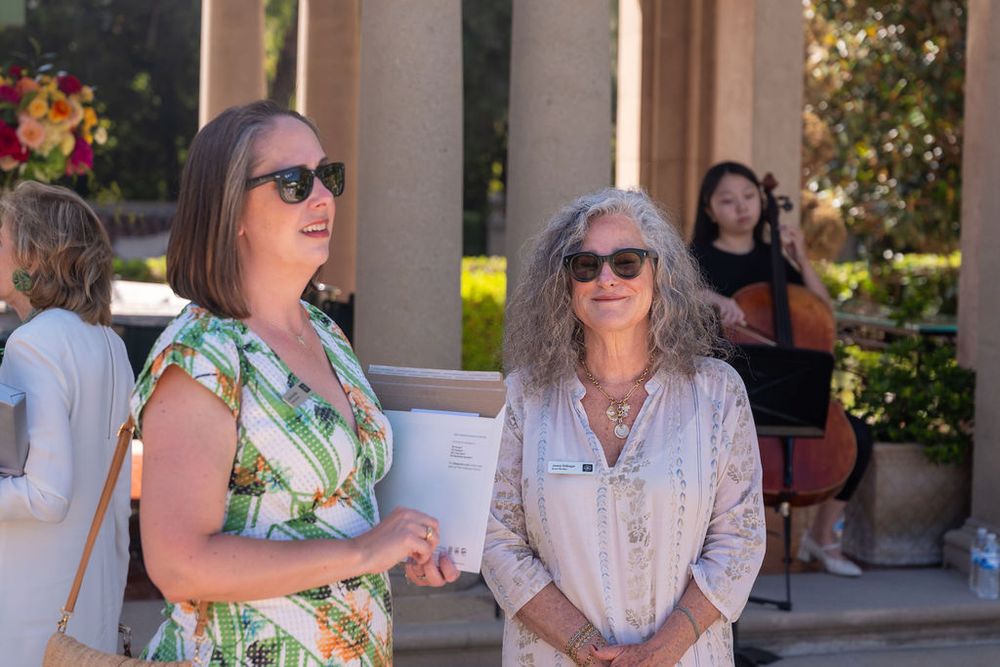 Two women in summer dresses chat outdoors at a garden event with a cellist performing in the background.