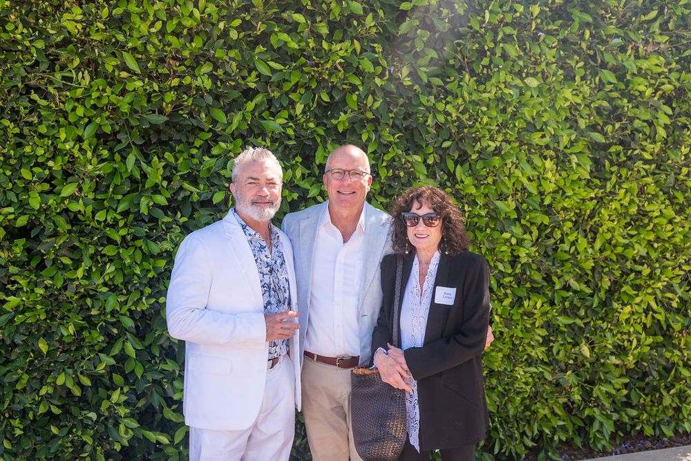 Three smiling adults posing together outdoors in front of a lush green hedge wall at a social event.
