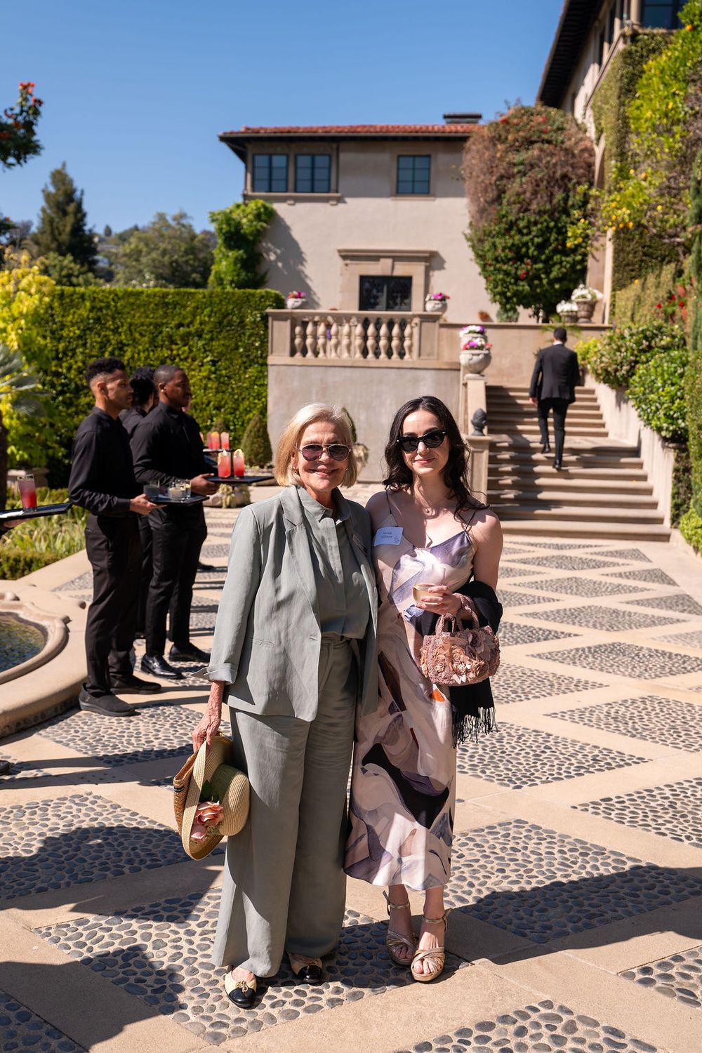 Two elegantly dressed women pose together in a sunlit courtyard of a luxury villa during an outdoor event.
