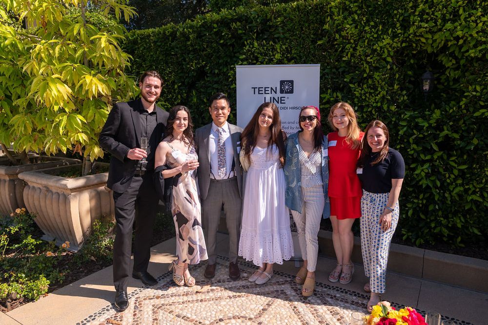 Group of seven young adults posing together outdoors at a Teen Line event, smiling in semi-formal attire.