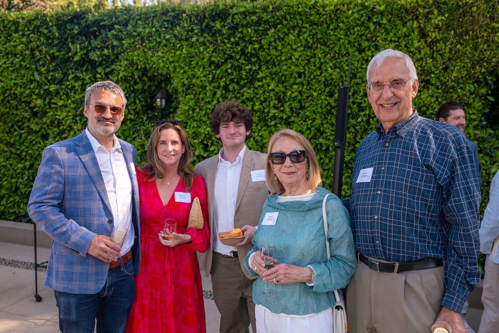 Five adults smiling and posing together outdoors at a social gathering near green hedges on a sunny day.