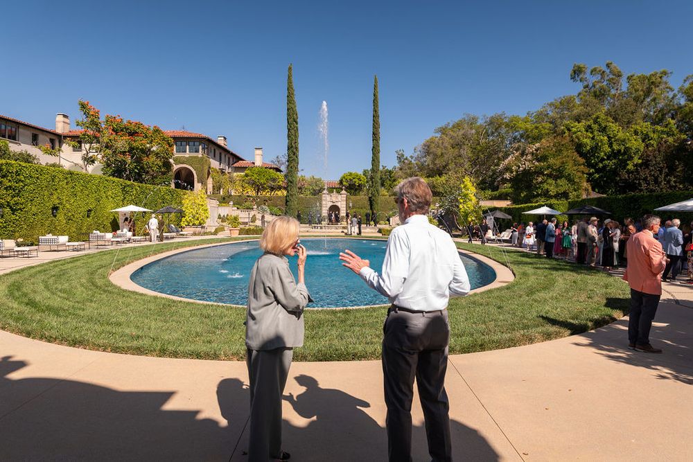 Two people overlooking an elegant estate garden with a circular fountain, cypress trees, and manicured lawns.