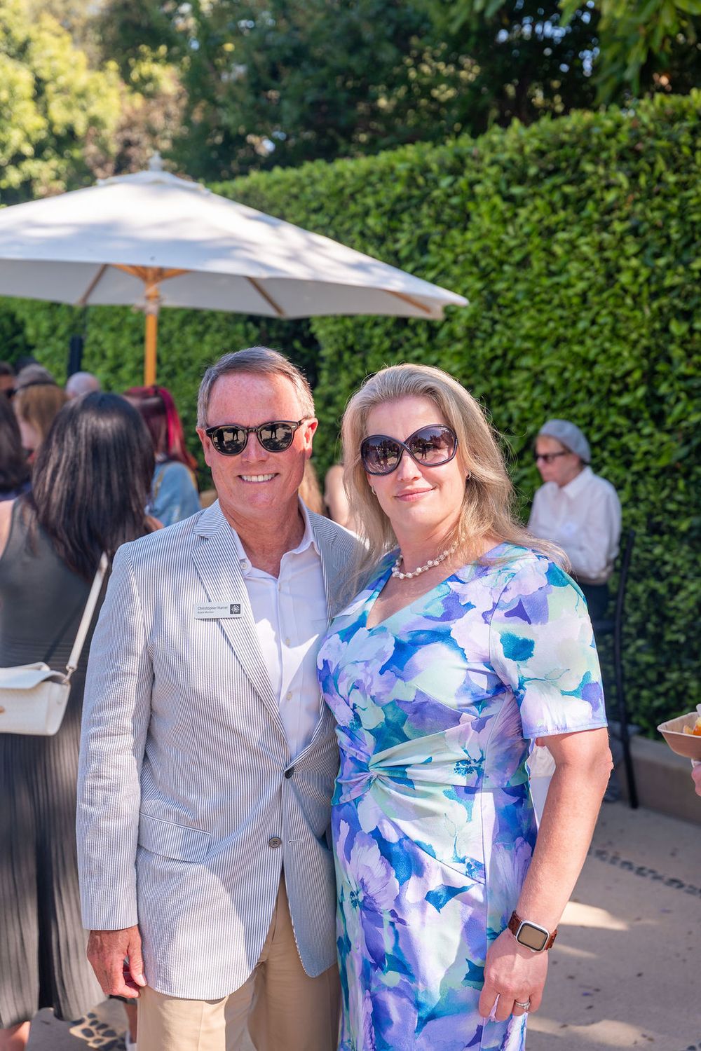 Elegant couple posing outdoors at a garden party, man in seersucker suit, woman in floral dress with pearl necklace.