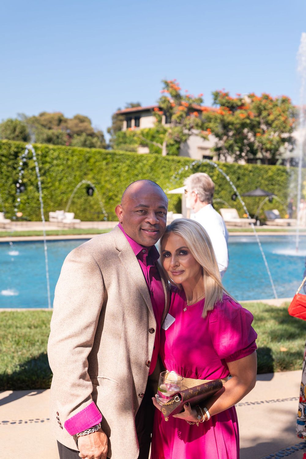Couple in pink and beige attire posing outdoors near a fountain at an elegant garden event on a sunny day.