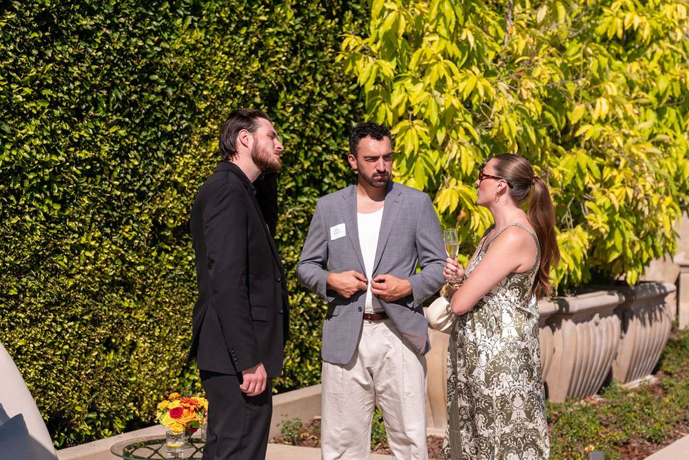 Three professionals networking outdoors at a social event, conversing in front of a lush green hedge wall.
