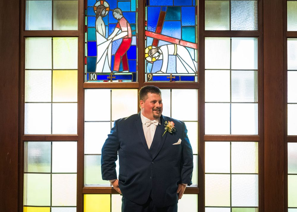 Groom in blue suit standing before ornate stained glass church window depicting religious scenes on his wedding day.