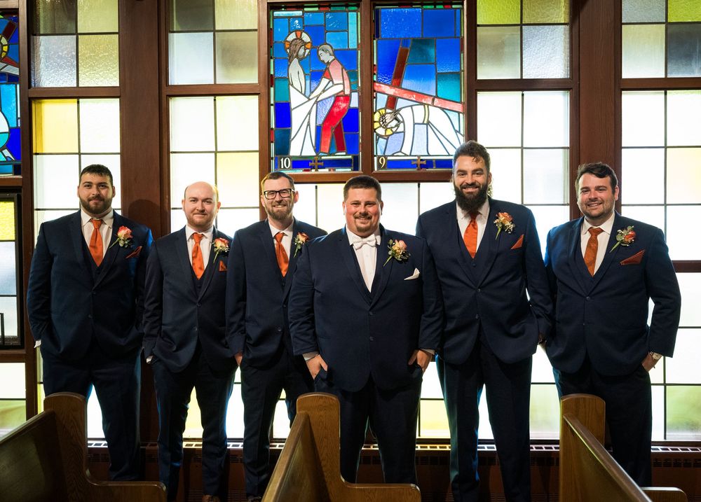 Six groomsmen in navy suits with orange boutonnieres pose in a church with colorful stained glass windows.