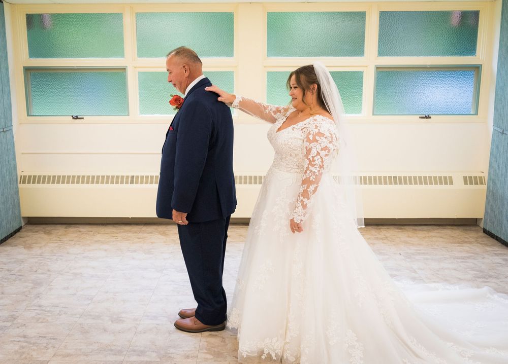 Bride in lace wedding gown adjusts groom's navy suit collar during wedding first look ceremony indoors.